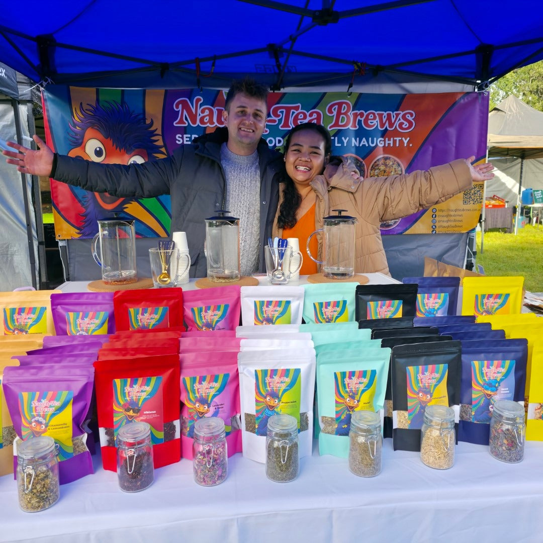 Two people standing behind a table with colorful tea packages and jars at a market stall. Naugh-Tea Brews Market Stall.
